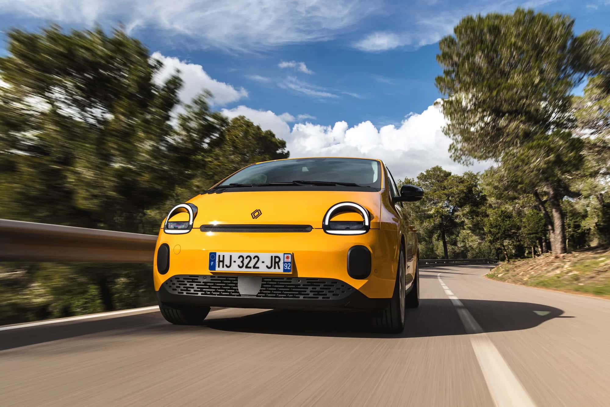 Renault Twingo E-Tech Techno 2026 en jaune Mango, vue de face en roulage sur route bordée de pins, dynamisme et signature lumineuse