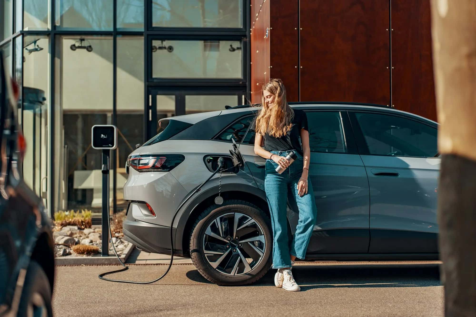 Une femme appuyée contre une voiture électrique grise en recharge devant un immeuble