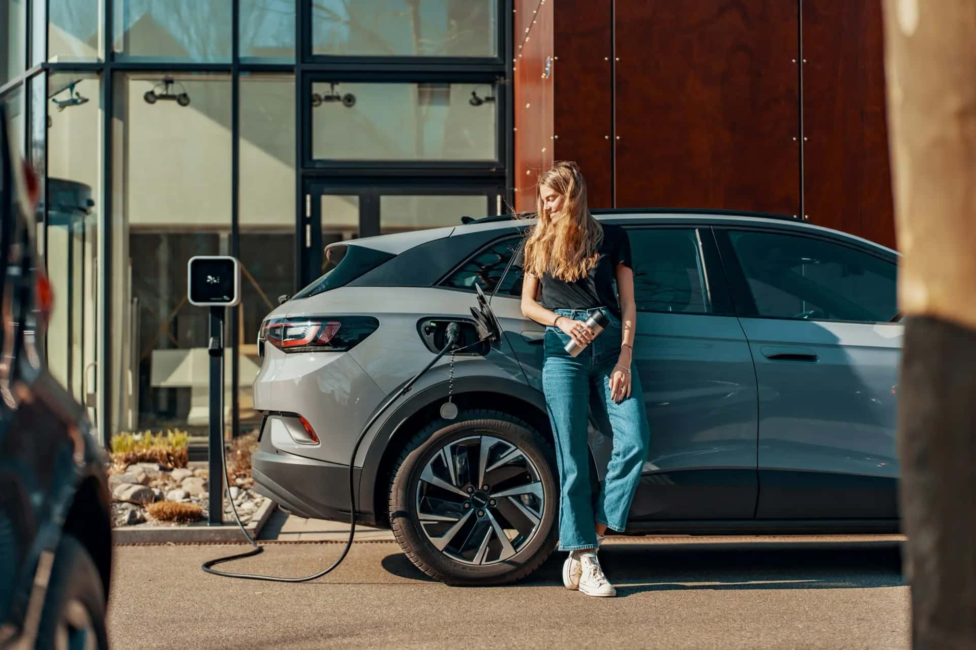 Une femme appuyée contre une voiture électrique grise en recharge devant un immeuble